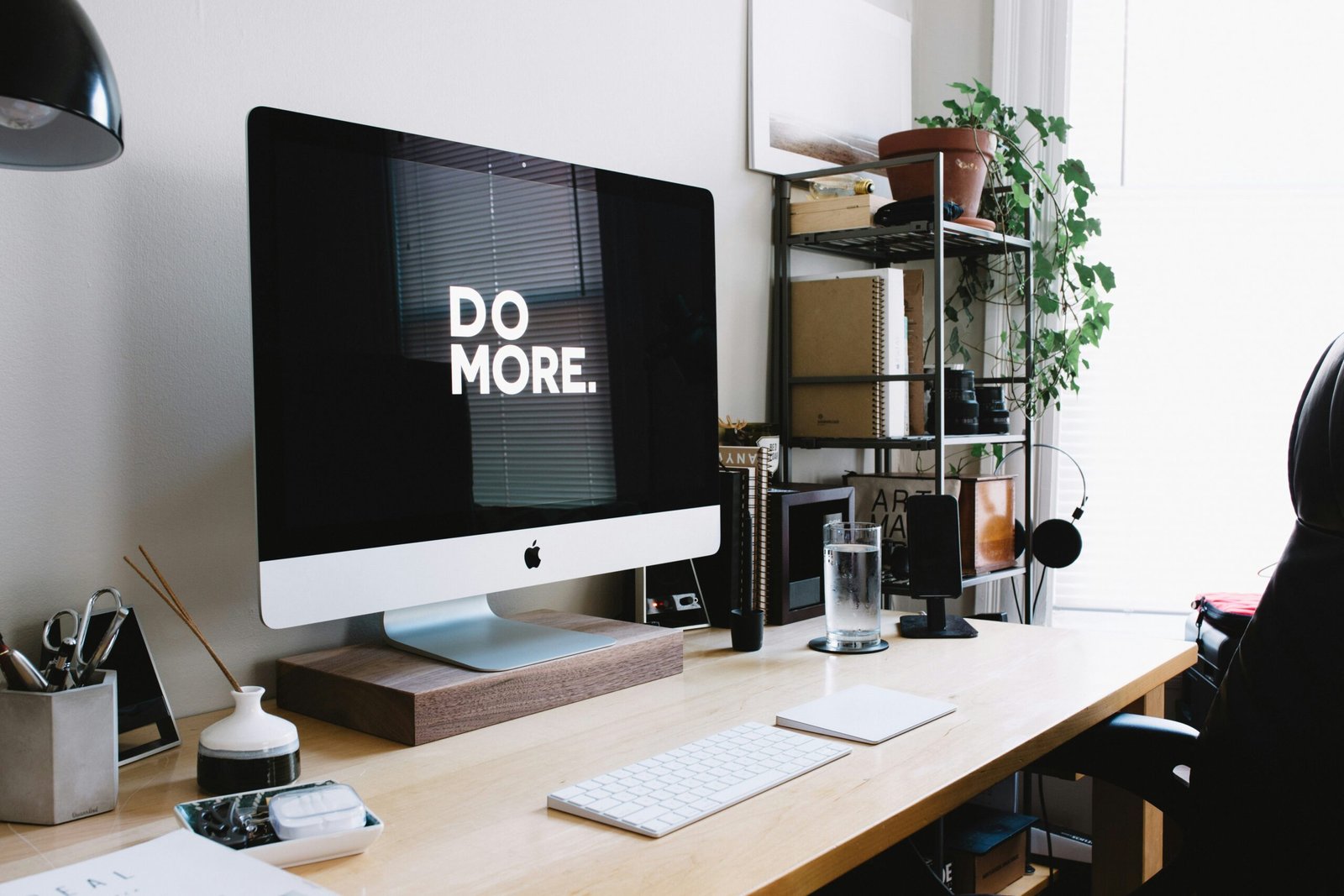 A modern workspace with an iMac displaying the words 'DO MORE.' on the screen, a wooden desk and neeatly arranged office supplies